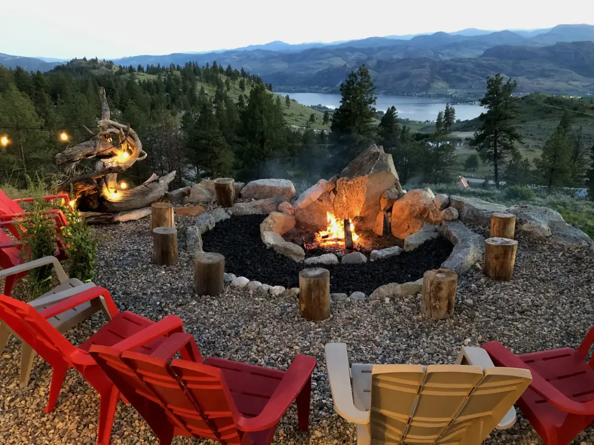 Vista Peak stone fire pit with colorful chairs, log stools, and driftwood art at dusk